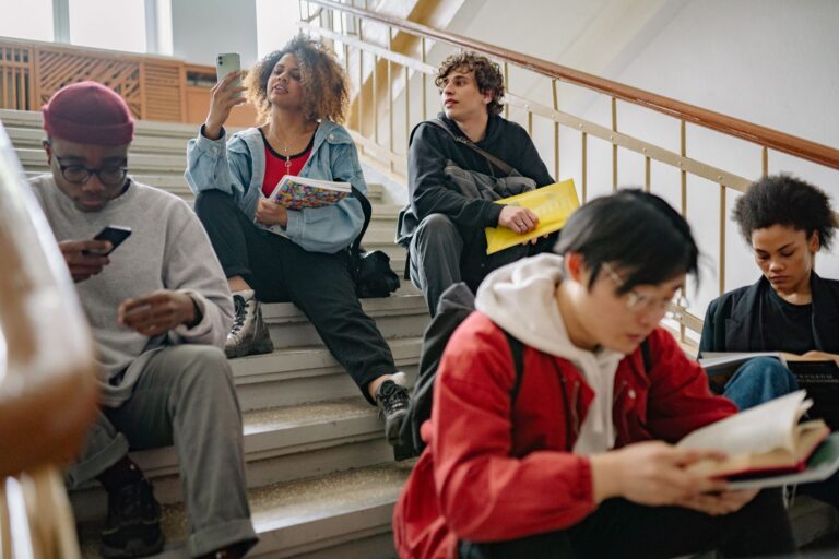 Studenci siedzą na schodach uczelni, uczą się i korzystają z telefonów